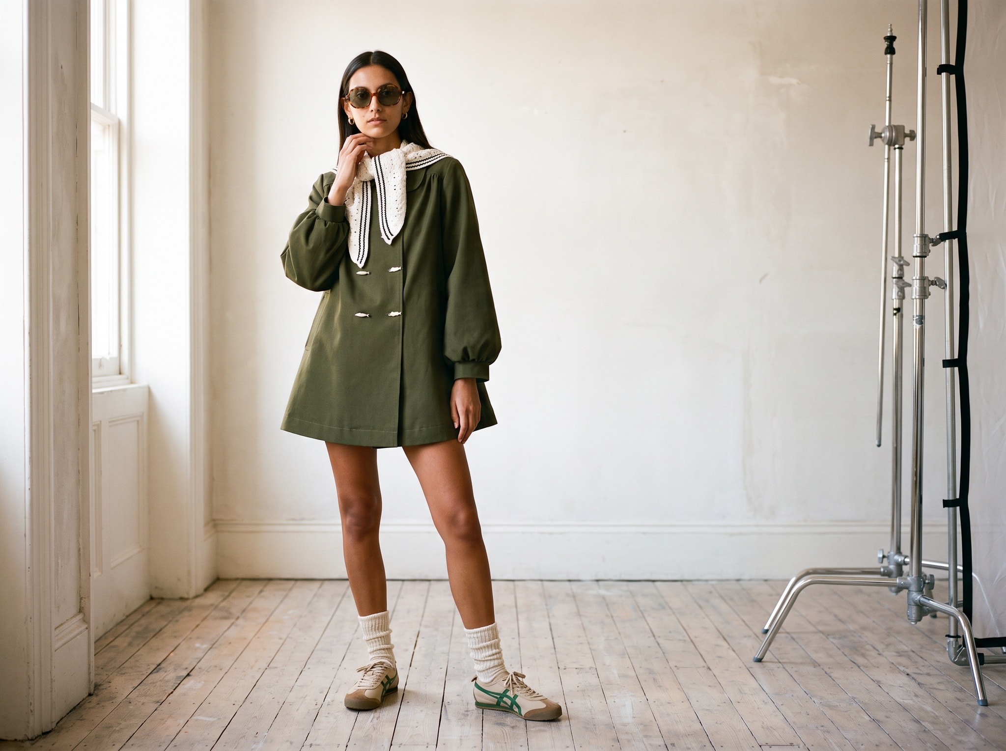 Inside the studio: a model in the caper-green coat standing in sunlit window light, a diffusion scrim and light stand visible mid-session