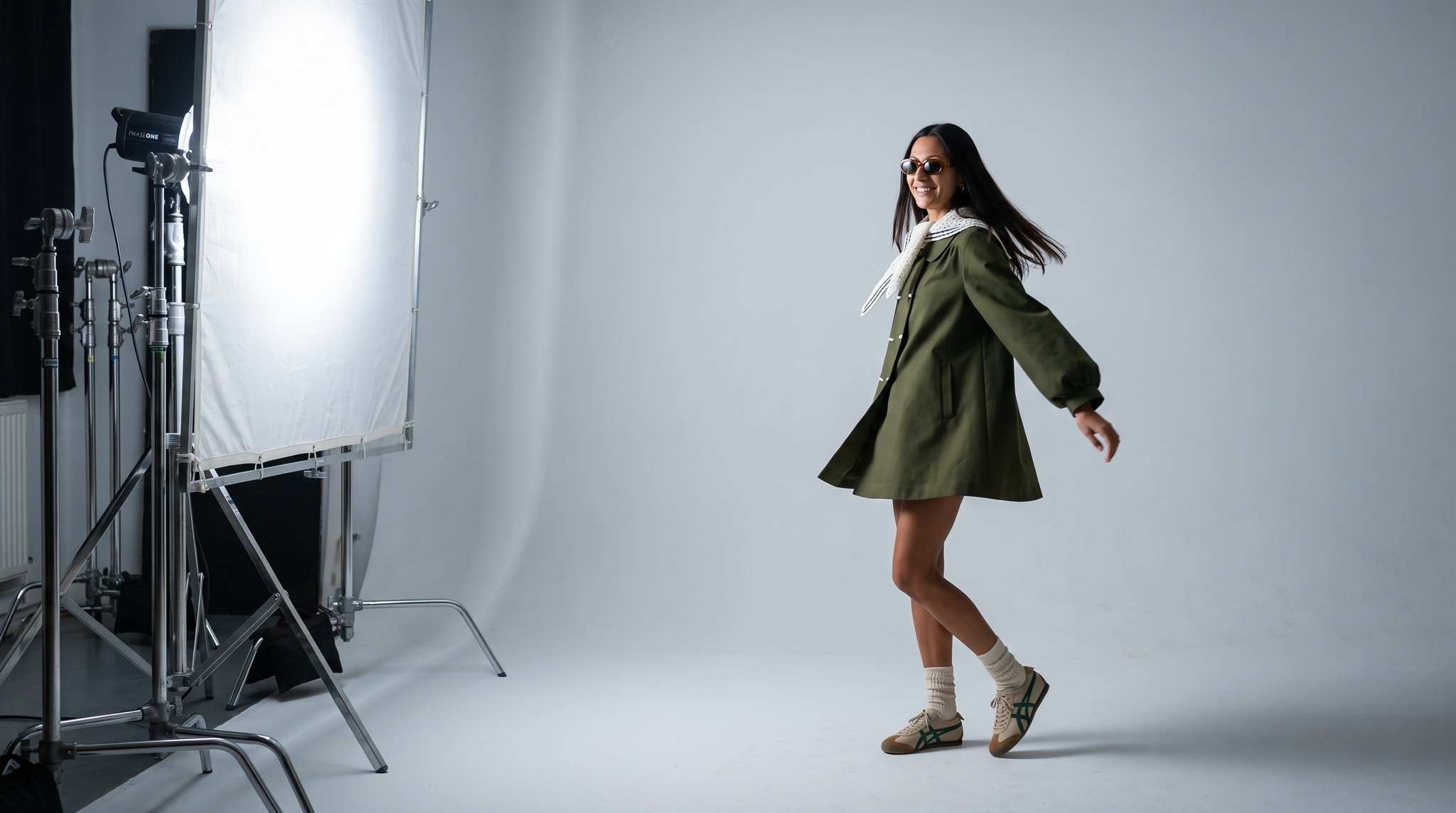 A model in the caper-green coat with a white crochet scarf standing on pale grey studio seamless, light stands and a diffusion frame at the left edge of the set