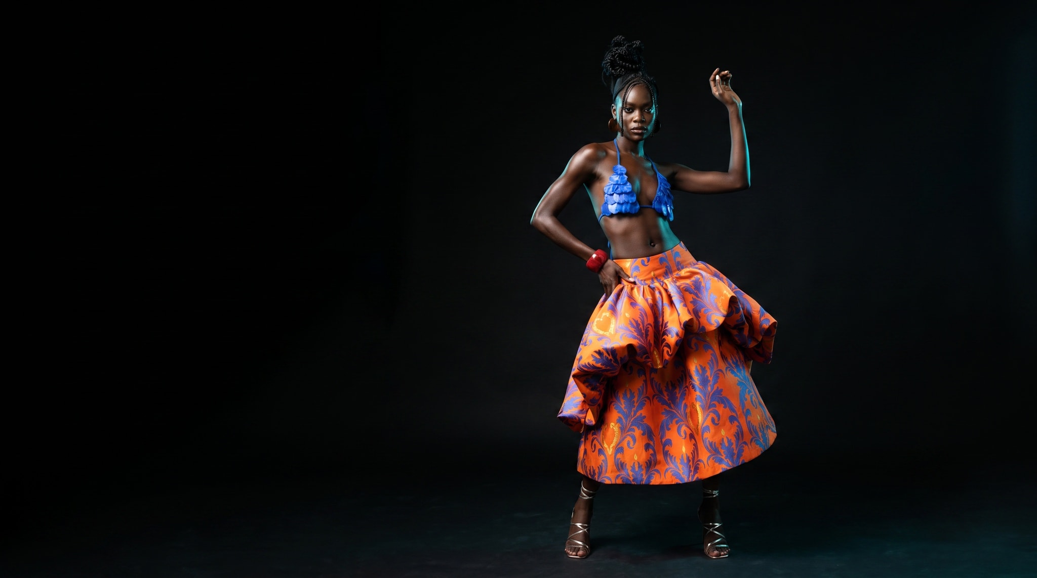 A dark editorial frame from the Ferendy studio: a model mid-movement in an orange brocade bubble skirt and blue halter, lit by a single beam against deep shadow