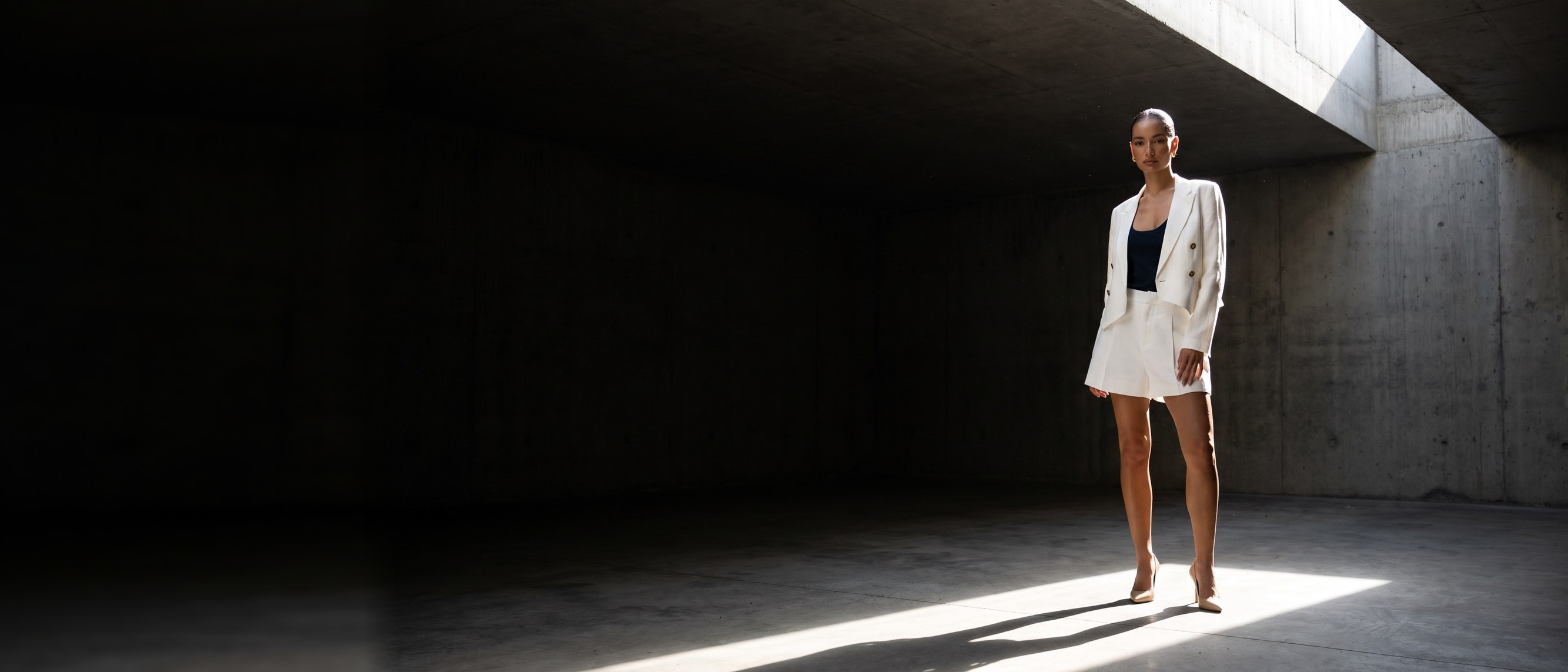 A model in an ivory linen suit standing in a hard shaft of daylight inside a dark brutalist concrete atrium, deep shadow filling the left of the frame
